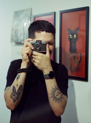 Scanned film photograph of a person (a man with short black hair and a mustache wearing a black t-shirt) looking
            through the viewfinder of an old camera at the photographer.